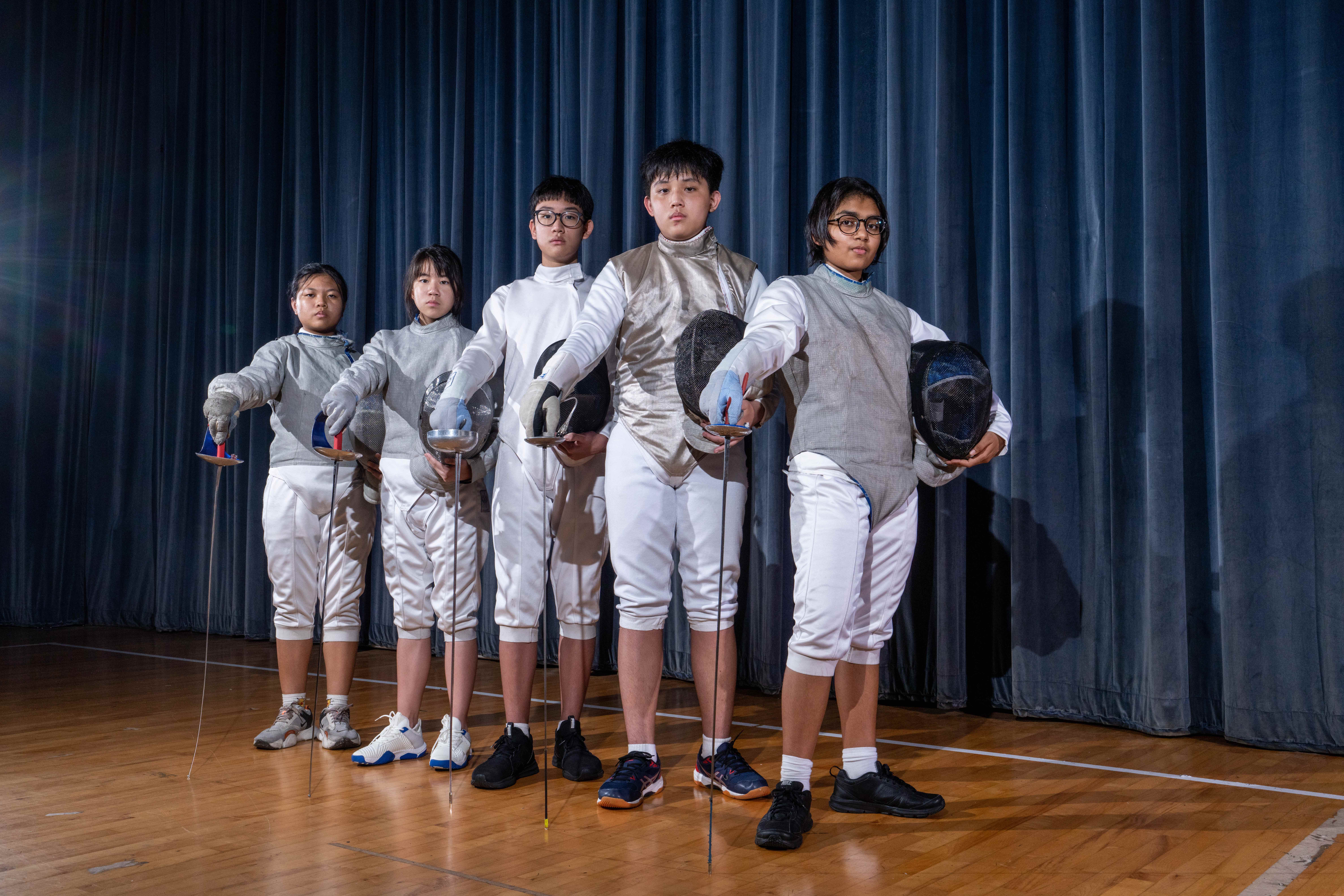 Five youth fencers in white gear pose with foils and masks on a gym floor before blue curtains.